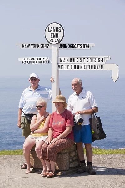 Friends on holiday having their photo taken at Lands End with the distance to the town where they live