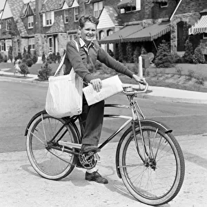 Paperboy on bicycle in suburban neighborhood, with sack over shoulder and folded newspaper in hand